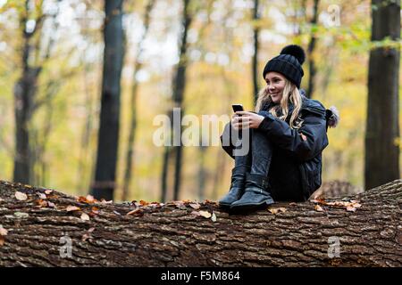 Mädchen mit Smartphone auf Baumstamm im herbstlichen Wald Stockfoto