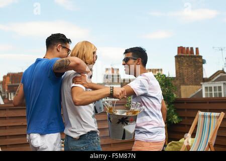 Drei männliche Freunde tragen Eiseimer mit Flaschenbier auf Party auf dem Dach Stockfoto