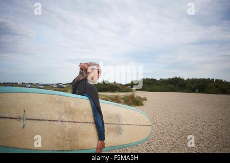 Ältere Frau zu Fuß entlang dem Strand, mit Surfbrett, Rückansicht Stockfoto