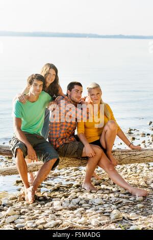 Freunde sitzen auf Treibholz neben Küste Blick in die Kamera Lächeln, Schondorf, Ammersee, Bayern, Deutschland Stockfoto