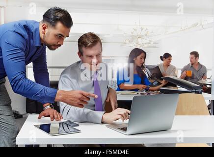 Zwei Geschäftsleute, Diskussion, mit Blick auf Laptop, im Hintergrund arbeitenden Kolleginnen und Kollegen Stockfoto