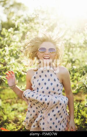 Junge Frau trägt ärmelloses Kleid und Sonnenbrille springen, Blick auf die Kamera zu Lächeln Stockfoto