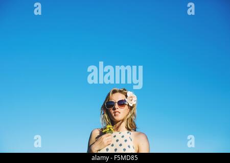 Niedrigen Winkel Blick der jungen Frau mit Sonnenbrille, Blumen im Haar halten Löwenzahn gegen blauen Himmel Stockfoto