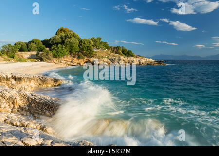 Eine Welle stürzt über die Felsen am Strand von Batariaand Kanoni, Kassiopi, Corfu. In der Ferne sehen die Berge von Albanien. Stockfoto