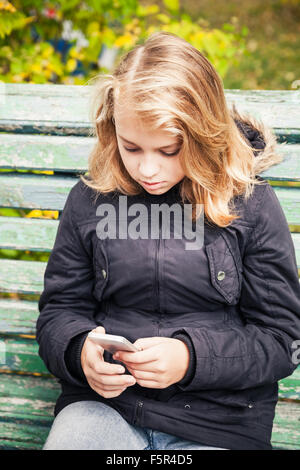 Schöne kaukasischen blonde Teenager-Mädchen in schwarzen Jacke sitzen auf grüne Bank mit Smartphone in Händen Stockfoto