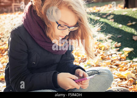 Niedliche kaukasischen blonde Teenager-Mädchen in Jeans und schwarze Jacke im Park sitzen und mit Smartphone, Outdoor-Herbst Porträt Stockfoto