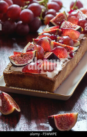 Hausgemachte rechteckige Torte mit roten Trauben, Feigen und Schlagsahne in weißen Keramikplatte über rote Holztisch. Dunkel rustikalen styl Stockfoto