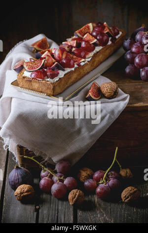 Hausgemachte rechteckige Torte mit roten Trauben, Feigen, Nüssen und Schlagsahne in weißen Keramikplatte auf hölzerne Truhe über alte woode Stockfoto