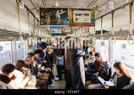 Japan, Osaka. Innenraum der Nankai Bahn überfüllten Pendlerzug mit Menschen stehen und sitzen Verweilen lesen Zeitungen und Telefone, Winter. Stockfoto