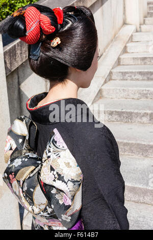 Japan, Kyoto. Frau mit schwarzen Kimono, im Sonnenlicht auf Stein Schritte stehen, entfernt mit Blick auf das Detail der Frisur und den Obi, den Bogen zurück zu zeigen. Stockfoto