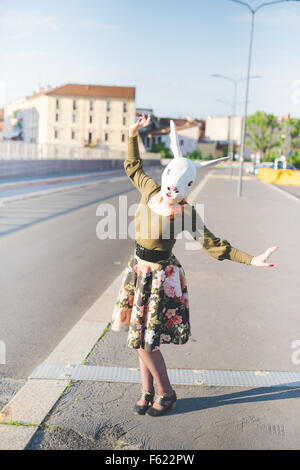 junge hübsche kaukasischen rothaarige Frau mit Hasenmaske, Tanz auf der Straße der Stadt, hält einen Streifen von ihren floralen Rock Stockfoto