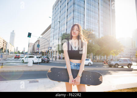 Knie-Figur des jungen schönen asiatischen lange braune glatte Haare Frau Skater posiert in der Stadt, in der Kamera suchen, nachdenklich - sportlich, nachdenklich, ernst-Konzept Stockfoto