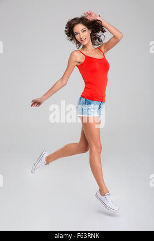 Glücklich lächelnd lockiges rotes Top, Jeans Shorts und weiße Turnschuhe im Sprung Mädchen in inspiriert Stockfoto