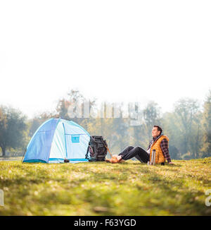 Entspannte junge Wanderer sitzen auf dem Rasen im Freien neben einem blauen Zelt an einem schönen Herbsttag schoss mit Tilt und shift Objektiv Stockfoto