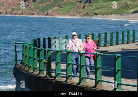 Nehmen Sie am Meer ein älteres Paar, Arm in Arm, einen erfrischenden Spaziergang entlang der Strandpromenade an einem hellen sonnigen Sommertag bei Whitbys, N Yorkshire, GB, UK. Stockfoto