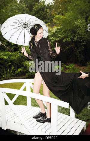Modebewusste Frau trägt schwarzes Kleid auf kleine Brücke stehend und weißen Schirm halten Stockfoto