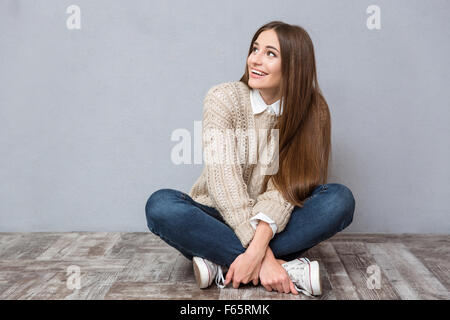 Happy begeistert junge Frau mit langen Haaren in Beige Pullover und Jeans sitzen auf Holzboden mit Beine gekreuzt und wegsehen Stockfoto