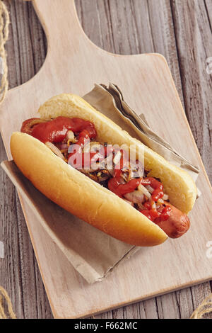 Hot Dog mit Zwiebeln, Ketchup und Senf auf einem weißen Schneidebrett auf einem Holztisch. Ketchup und Senf in zwei transparente Schüssel Stockfoto