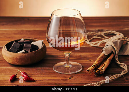 Glas Schnaps auf einem alten Holztisch mit einer Schüssel mit Schokolade, Zimt Stiks und Chili Paprika. Warmen Hintergrund. Stockfoto