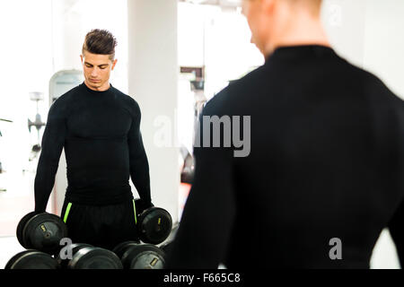 Muskulöser Jüngling, trainieren Sie im Fitnessraum und heben von Gewichten mit seiner Reflexion in einem Spiegel zeigen Stockfoto