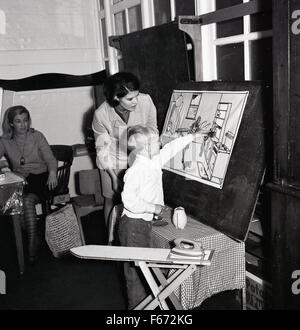 1950er Jahre historische, kleine Jungen im Kindergarten mit Lehrerin Dinge auf einer Zeichnung einer Küche auf. Stockfoto