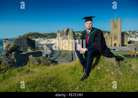 Männliche Aberystwyth Universitätsstudent tragen traditionelle Kappe und Kleid und Mörtel Board am Abschlusstag, sitzen im Freien mit der Hochschule und die Stadt hinter ihm, Wales UK Stockfoto