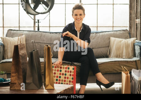 Mühelos stilvolle Brünette Frau mit bunten Einkaufstüten auf Sofa im modernen Loft-Wohnung sitzen. Gute shopping-Tour macht jede Frau glücklich. Luxuriöses Leben-Konzept Stockfoto