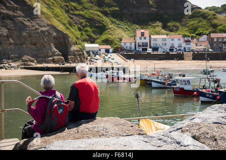 Paar, sitzen in der Sommersonne, bewundern Sie die Aussicht auf Angelboote/Fischerboote vertäut im Hafen & malerischen Küsten Dorf von Staithes, North Yorkshire, UK. Stockfoto