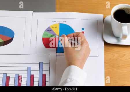 Frau Hand mit Stift und zeigt im Kreisdiagramm mit gedruckten Diagrammen, teilweise Laptop und Kaffee auf Desktop. Stockfoto