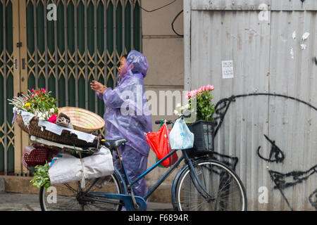 Vietnamesische Dame in Kunststoff Mac verkauft Blumen auf der Straße vom Rad in Hanoi Altstadt, Vietnam Stockfoto