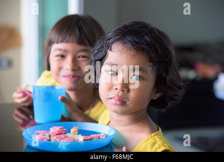 Zwei Thai-Mädchen, 3 und 7 Jahre alt, trinken Milch und Brot mit Salami zu essen. Beide Mädels haben einen sichtbaren Milch Schnurrbart. Stockfoto