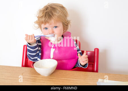 1 Jahr altes Baby Essen Joghurt messily mit einem Löffel, England, UK Stockfoto