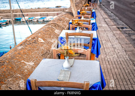 Esstische im italienischen Restaurant auf braune steinernen Brüstung auf dem Meer Bucht von Gallipoli (Le) in Italien an einem sonnigen Sommertag: Kunststoff gefälschte gelbe Blumen mit großen Blüten und grünen Blättern in weiße Vase, weiß und blau karierte Servietten und Tischdecke auf Elec Stockfoto