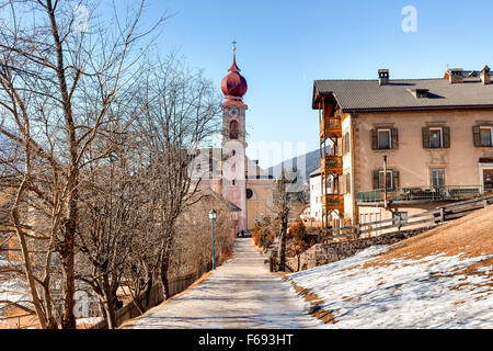 Der Luis Trenker Promenade führt bis St. Ulrich, italienisches Dorf in Dolomiten Alpen: typische Häuser, Gebäude und die roten Glockenturm mit Uhr Pfarrkirche St. Ulrich mit schneebedeckten Bergen und grünen Koniferen im Hintergrund Stockfoto