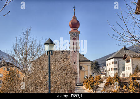 Der Luis Trenker Promenade führt bis St. Ulrich, italienisches Dorf in Dolomiten Alpen: typische Häuser, Gebäude und die roten Glockenturm mit Uhr Pfarrkirche St. Ulrich mit schneebedeckten Bergen und grünen Koniferen im Hintergrund Stockfoto