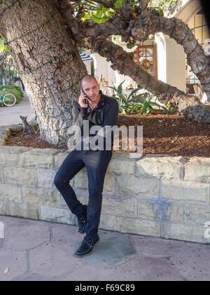 Ein 20-30 Jahre alten Mann in schwarz gekleidet sieht geradeaus wie er auf seinem Handy in Santa Barbara, Kalifornien spricht. Stockfoto