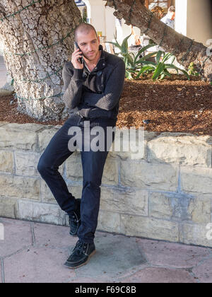 Ein 20-30 Jahre alten Mann in schwarz gekleidet sieht geradeaus wie er auf seinem Handy in Santa Barbara, Kalifornien spricht. Stockfoto