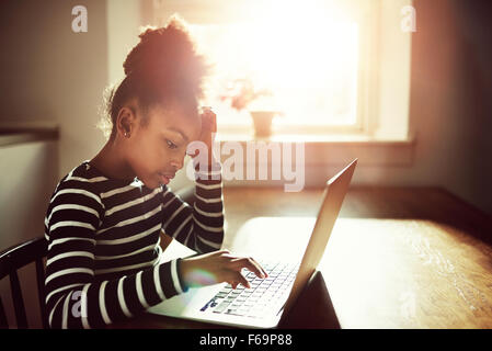 junge schwarze Mädchen arbeiten auf ihre Hausaufgaben zu Hause Eingabe von Informationen auf einem Laptopcomputer, gegen eine helle Sonne glühen Seitenansicht Stockfoto