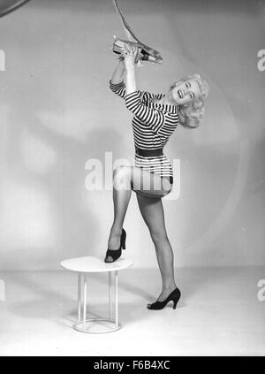 Dieses Schwarzweiß-Foto aus dem Jahr 1955 zeigt das Modell Rita Fisher, das in einer auffälligen Pose für eine Werbekampagne aufgenommen wurde. Das Bild konzentriert sich auf ihr glamouröses Styling und die Konsumkultur der 1950er Jahre mit Schwerpunkt auf Kleidung, Kosmetik und Mode. Stockfoto