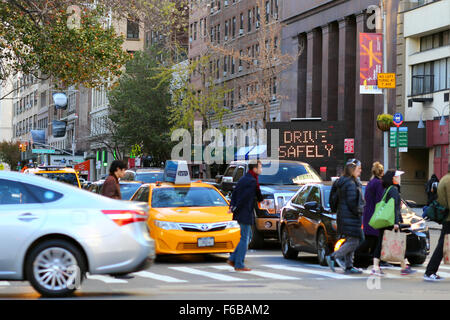 Ein elektronisches Schild blinkt Fahren Sie sicher in der Nähe einer Kreuzung von New York City, während Fußgänger über eine Straße neben Autos gehen, die auf dem Fußgängerübergang angehalten wurden. Stockfoto