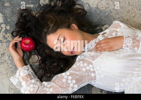 Atemberaubende junge Frau im weißen Spitzenkleid auf dem Boden liegend, mit einem roten Apfel Stockfoto