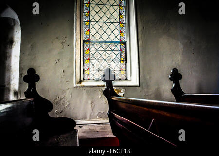 Ein schwach beleuchteten Innenraum aus einer Kirche Glasfenster mit geschnitzten Kreuze dekorativ an den Enden jedes Holz Pew Kunst gesetzt Stockfoto