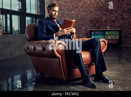 Junge hübsche Geschäftsmann mit Bart im schwarzen Anzug sitzen auf Stuhl Buch zu lesen. Konzentrieren Sie sich auf Gesicht. Stockfoto