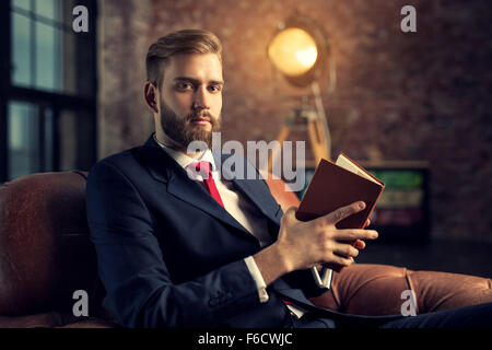 Junge hübsche Geschäftsmann mit Bart im schwarzen Anzug sitzen auf Stuhl Buch zu lesen. Stockfoto