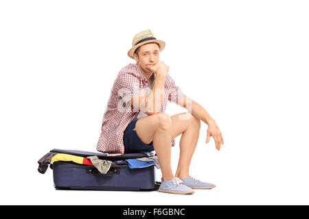 Traurige männliche Touristen sitzen auf Tasche voller Kleidung und schaut in die Kamera, die isoliert auf weißem Hintergrund Stockfoto