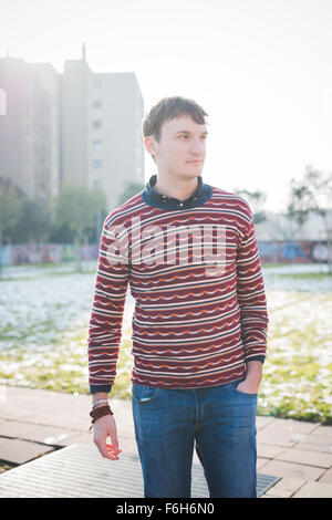 Knie-Figur des schönen jungen Mann mit Blick auf Links in der Stadt, trägt blaue Jeans und ein rot gestreiften Pullover, Hände in der Tasche- Stockfoto