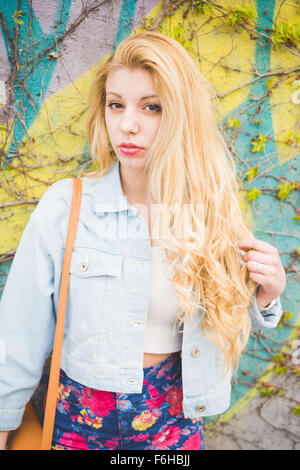 Junge hübsche blonde kaukasische Mode Mädchen posiert stützte sich auf einer Wand in die Vororte der Stadt ihr Haar berühren, suchen in der Kamera - Konzept von Jugend und unbeschwert Stockfoto