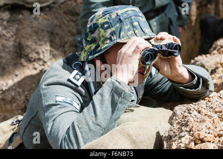 WW2re-enactment. Deutsche Wehrmacht Officer in Graben auf der Suche über den oberen durch ein Fernglas im hellen Sonnenschein verdeckt. Camouflage Helm trägt. Stockfoto