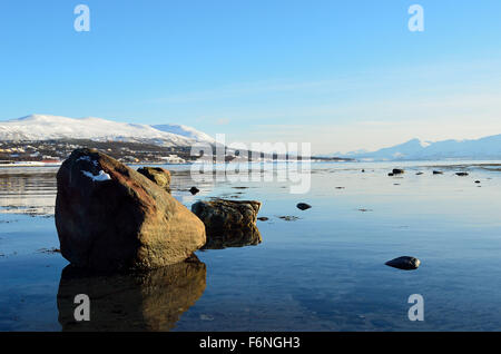 massiven Felsblock auf ruhiger See Fjord bei Sonnenschein mit verschneiten Hintergrund und Tromso Stadtinsel Stockfoto