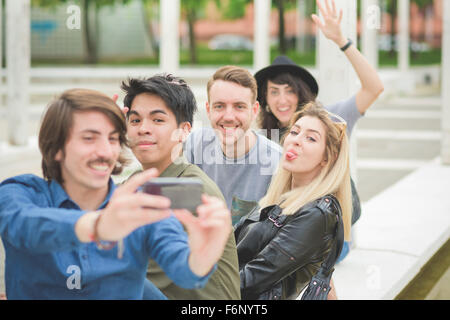 Gruppe junger multiethnischen Freunde auf einer kleinen Mauer sitzend, miteinander zu reden, per Smartphone, wobei eine Selfie, haben Stockfoto